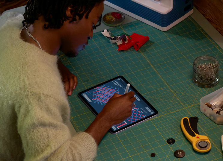 Eine Person, die mit dem Apple Pencil auf dem iPad zeichnet und an einer grünen Schneidematte mit Nähzubehör sitzt.